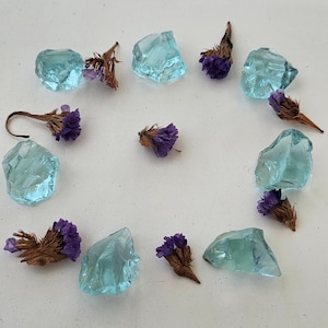 Puede incluir: Una colección de pequeños trozos de vidrio azul claro dispuestos en círculo sobre una superficie blanca. Flores secas moradas están dispersas alrededor de los trozos de vidrio.