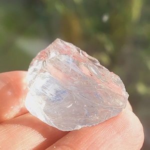 May include: A clear, faceted crystal with a rough, irregular shape. The crystal is held in a hand against a blurred background of green foliage.