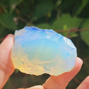 Puede incluir: Un gran cristal de ópalo iridiscente, blanco, azul y amarillo. El cristal es rugoso y tiene una forma natural irregular.