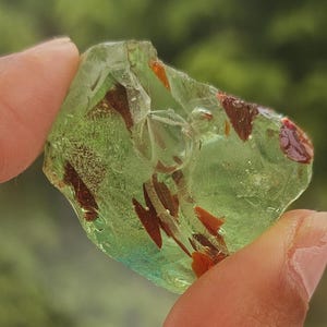 May include: A piece of green resin with embedded brown and red accents is held up against a blurred green background. The resin is translucent, allowing light to pass through, and is held between fingers.