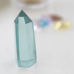 May include: A light blue crystal point stands upright on a white surface. The crystal is faceted with a pointed top. In the background, a blurred glass and other small objects are visible.