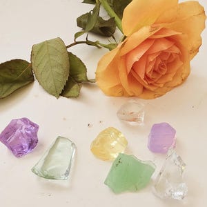 Peut inclure: Une rose orange vif avec des feuilles vertes et une collection de pierres de verre colorées de forme irrégulière. Les pierres sont dans les tons de violet, jaune, vert et transparent. La rose est en pleine floraison.