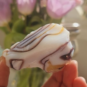 Puede incluir: Una piedra de ágata rayada de blanco, morado y dorado con una superficie lisa y pulida.