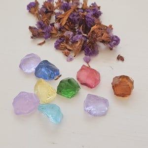 Peut inclure: Une collection de petites pierres de verre colorées dans des tons de violet, bleu, vert, jaune, rose et orange. Les pierres sont éparpillées sur une surface blanche, avec un tas de fleurs violettes séchées.