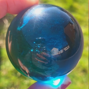 May include: A teal blue glass sphere with a reflective surface. The sphere is being held in a hand.