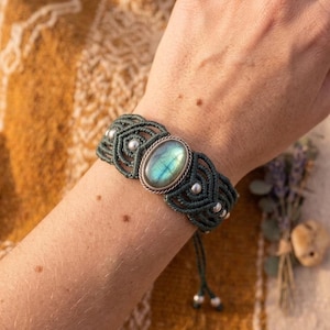 Puede incluir: Una pulsera de macramé verde oscuro con una piedra preciosa de labradorita ovalada central con brillo metálico, acentuada por pequeñas cuentas plateadas tejidas en el patrón de cordón, terminada con dos extremos de cordón colgantes con cuentas plateadas.