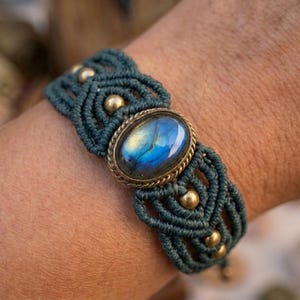 May include: A dark teal macramé bracelet featuring a central oval labradorite gemstone set in a brass-toned metal frame. The woven cord design incorporates small, round, metallic brass-coloured beads arranged symmetrically along the band.