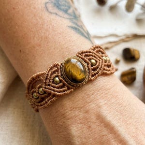Könnte beinhalten: Ein Makramee-Armband mit einem zentralen, ovalen Tigerauge-Edelstein in einer braunen Kordelfassung. Das Armband ist mit kleinen Metallperlen verziert, die in das komplexe Knotenmuster eingearbeitet sind und ein strukturiertes Band bilden.