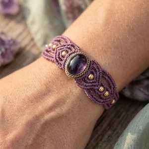 Peut inclure: Un bracelet en macramé avec une pierre précieuse améthyste violette de forme ovale sertie dans un cadre en métal doré. Le bracelet est composé d'un cordon violet tissé, orné de petites perles métalliques rondes de couleur dorée.