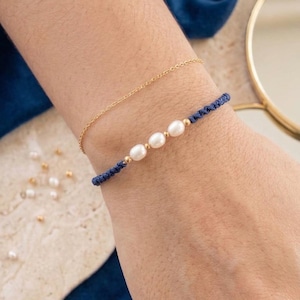 Peut inclure: Un bracelet en chaîne dorée fine associé à un bracelet en cordon bleu marine orné de trois perles blanches ovales séparées par de petites perles dorées. Le cordon bleu marine présente un tissage texturé et la chaîne dorée est fine.