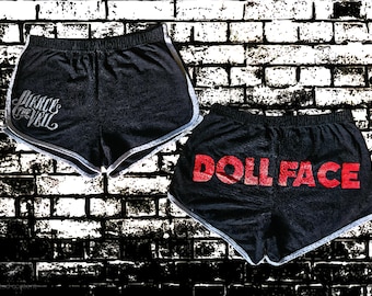 Puede incluir: Dos pares de pantalones cortos deportivos negros con ribete blanco. Uno presenta el texto "Pierce the Veil" en plata, y el otro muestra "DOLLFACE" en letras rojas.