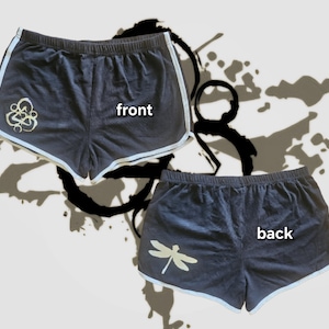 May include: Dark gray athletic shorts with white trim. The front of the shorts features a gold-colored abstract design, and the back displays a gold dragonfly. The word "front" is printed on the front, and "back" is printed on the back.