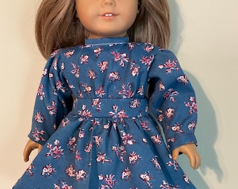 Könnte beinhalten: Eine Puppe mit schulterlangem hellbraunem Haar und blauen Augen, bekleidet mit einem langärmeligen blauen Kleid mit rosa-weißem Blumenmuster, einem weißen Unterrock mit Spitzenbesatz und dunkelbraunen Schnürstiefeln mit schwarzen Schnürsenkeln.