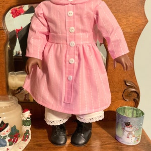 May include: A pink and white striped dress with a white collar and buttons. The dress is worn by a doll with dark skin and black hair. The doll is wearing black shoes and white lace-trimmed underwear.
