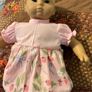 Peut inclure: Une poupée au teint clair vêtue d'une robe à manches courtes. La robe possède un corsage rose pâle uni et une jupe froncée ornée d'un motif répétitif de fraises rouges, de feuilles vertes, de fleurs blanches et de petits nœuds bleus sur fond rose.