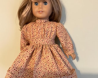 Puede incluir: Una muñeca con un vestido de manga larga con estampado floral en tonos rojos, azules y amarillos. El vestido tiene una cintura fruncida y un ribete de encaje blanco. La muñeca lleva calcetines blancos y zapatos negros.