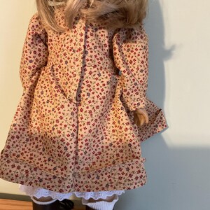 K&ouml;nnte beinhalten: Eine Puppe, die ein beigefarbenes Kleid mit Blumenmuster und braune Stiefel tr&auml;gt. Das Kleid hat einen wei&szlig;en Spitzenbesatz am Saum.