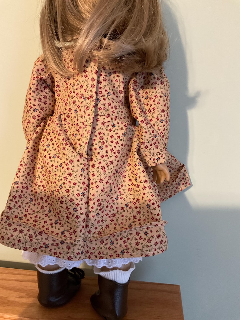 K&ouml;nnte beinhalten: Eine Puppe, die ein beigefarbenes Kleid mit Blumenmuster und braune Stiefel tr&auml;gt. Das Kleid hat einen wei&szlig;en Spitzenbesatz am Saum.