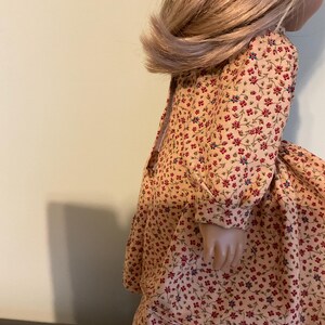 K&ouml;nnte beinhalten: Eine Puppe tr&auml;gt ein lang&auml;rmeliges Kleid mit einem Blumendruck in Rot-, Gelb- und Blaut&ouml;nen. Das Kleid hat einen gerafften Rock und ein tailliertes Oberteil.