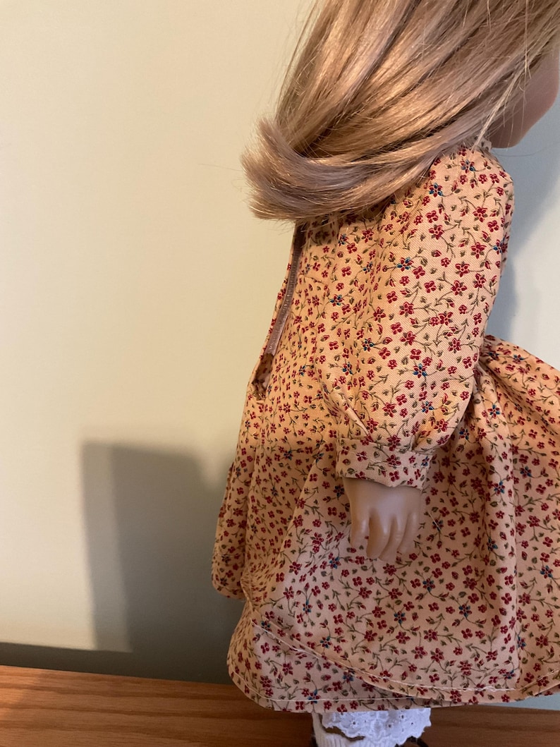 K&ouml;nnte beinhalten: Eine Puppe tr&auml;gt ein lang&auml;rmeliges Kleid mit einem Blumendruck in Rot-, Gelb- und Blaut&ouml;nen. Das Kleid hat einen gerafften Rock und ein tailliertes Oberteil.