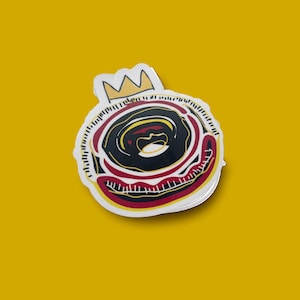 May include: A sticker of a donut with a crown on top. The donut is red, black, and yellow. The crown is gold.