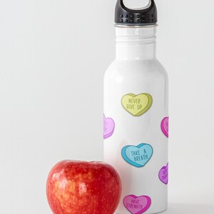 May include: A white water bottle with a black cap and colorful heart-shaped graphics with motivational phrases. A red apple sits next to the bottle. The phrases include "Never Give Up" and "Take a Breath".