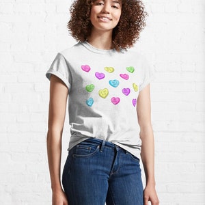 May include: A light gray t-shirt featuring colorful heart-shaped candies with various phrases. The candies are in shades of pink, yellow, green, and blue. The shirt is paired with blue jeans.