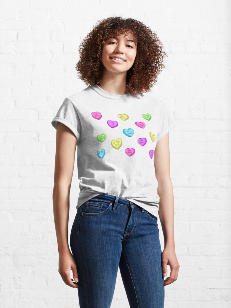May include: A light gray t-shirt featuring colorful heart-shaped candies with various phrases. The candies are in shades of pink, yellow, green, and blue. The shirt is paired with blue jeans.