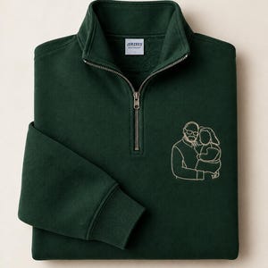 Könnte beinhalten: Ein dunkelwaldgrünes Sweatshirt mit Viertelreißverschluss, silberfarbenem Metallreißverschluss und einem weißen Etikett mit der Aufschrift JERZEES. Auf der Brust befindet sich eine helle Linienstickerei, die einen Erwachsenen mit einem Kind zeigt.