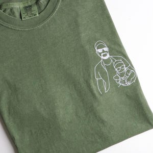 Peut inclure: T-shirt Comfort Colors vert olive avec une illustration blanche d'une personne tenant un enfant. Le t-shirt a un col rond et des manches courtes. L'étiquette indique XL - TG - EG.
