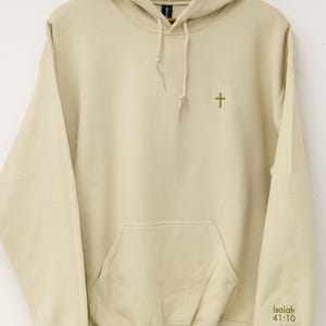 May include: A beige hoodie with a front pocket and a drawstring hood. A small green cross is embroidered on the chest, and the text "Isaiah 41:10" is printed on the right sleeve. The hoodie is hanging on a wooden hanger.