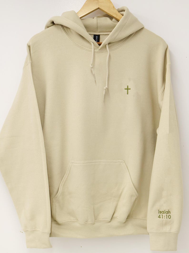 May include: A beige hoodie with a front pocket and a drawstring hood. A small green cross is embroidered on the chest, and the text "Isaiah 41:10" is printed on the right sleeve. The hoodie is hanging on a wooden hanger.
