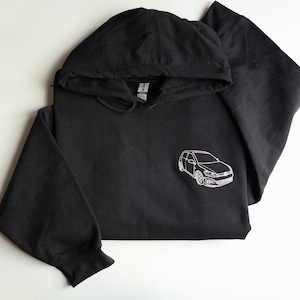 May include: Black hoodie with a white embroidered car graphic. The sweatshirt features a drawstring hood and long sleeves. The car graphic is a detailed outline of a hatchback, a popular car style in the UK.