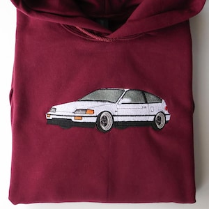 May include: Maroon-coloured hoodie featuring a white embroidered car design. The car is a coupe with a hatchback, with a white body, black tyres, and silver rims. The hoodie has a drawstring and a front pocket.