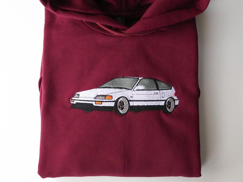May include: Maroon-coloured hoodie featuring a white embroidered car design. The car is a coupe with a hatchback, with a white body, black tyres, and silver rims. The hoodie has a drawstring and a front pocket.