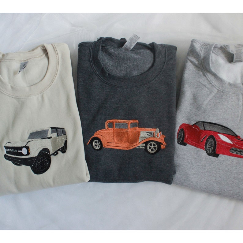 May include: Three sweatshirts with embroidered car designs. A cream sweatshirt features a white SUV. A dark grey sweatshirt has an orange vintage car. A light grey sweatshirt displays a red sports car.