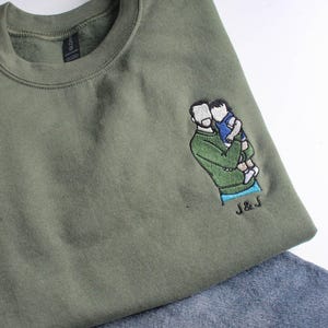 Puede incluir: Sudadera verde oliva con un dise&ntilde;o bordado de una persona sosteniendo a un ni&ntilde;o. El dise&ntilde;o es en tonos verdes, azules y blancos. Las iniciales "J & J" est&aacute;n bordadas debajo del dise&ntilde;o. La sudadera se combina con mezclilla azul.