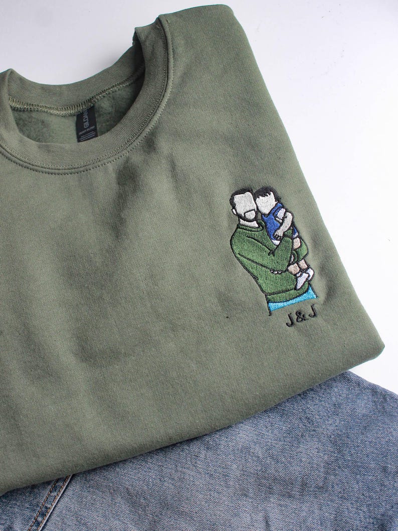 Puede incluir: Sudadera verde oliva con un dise&ntilde;o bordado de una persona sosteniendo a un ni&ntilde;o. El dise&ntilde;o es en tonos verdes, azules y blancos. Las iniciales "J & J" est&aacute;n bordadas debajo del dise&ntilde;o. La sudadera se combina con mezclilla azul.