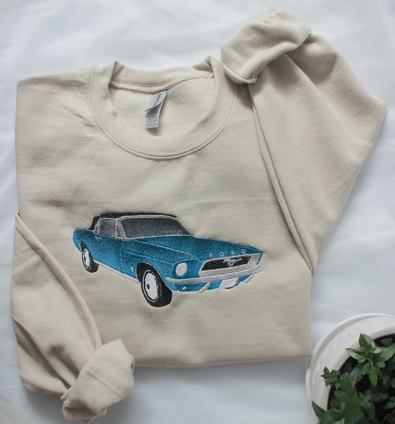 May include: A beige sweatshirt with an embroidered design of a blue classic car. The car has a black roof and silver details, with the word "FORD" visible. The sweatshirt is made of a soft material and has a crew neck.