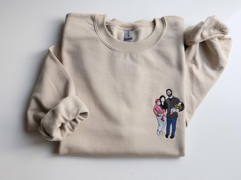 Puede incluir: Sudadera beige de cuello redondo con un retrato familiar bordado en la parte delantera. El retrato representa a una pareja con un beb&eacute; y un ni&ntilde;o. La sudadera est&aacute; doblada, con una manga visible.