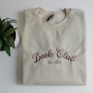 May include: A beige sweatshirt with the embroidered text "Book Club Est. 2021" in a brown script font. The sweatshirt is folded neatly on a white surface, with a potted plant visible in the corner.