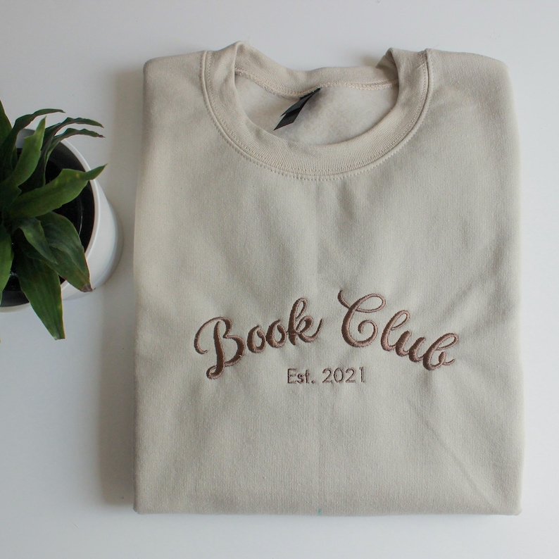 May include: A beige sweatshirt with the embroidered text "Book Club Est. 2021" in a brown script font. The sweatshirt is folded neatly on a white surface, with a potted plant visible in the corner.