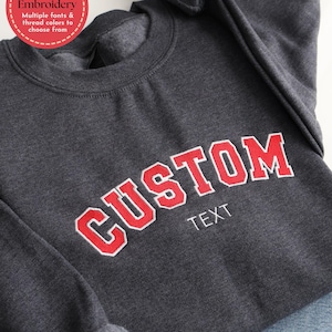 May include: Dark grey sweatshirt with the word "CUSTOM" embroidered in red with a white outline. The word "TEXT" is embroidered below. A red circle with white text reads "Custom Embroidery, Multiple fonts & thread colours to choose from."
