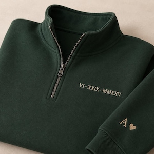 Pode incluir: Uma camisola verde-floresta escura com fecho de correr de um quarto, apresentando a data em numerais romanos VI • XXIX • MMXXV bordada a fio dourado no peito e a letra A seguida de um pequeno coração bordados a ouro no punho.
