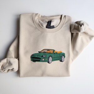 May include: A cream-coloured sweatshirt featuring an embroidered green convertible car. The car has a tan interior and silver wheels. The sweatshirt has a crew neck and long sleeves. The car design is centered on the front.