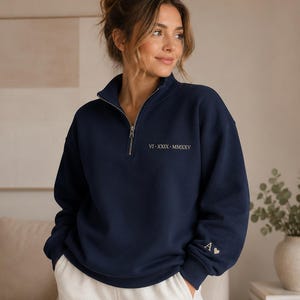 May include: A navy blue quarter-zip sweatshirt featuring a silver-toned zip. The chest displays the Roman numeral date VI-XXIX-MMXXV in light-coloured text, while the left sleeve cuff features the letter A followed by a small heart symbol.
