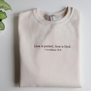 May include: A cream-coloured sweatshirt with the text "Love is patient, love is kind" and the reference "1 Corinthians 13:4" in a brown font. The sweatshirt is folded neatly on a white surface.