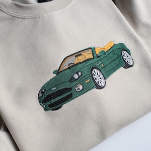 May include: A cream-coloured sweatshirt with an embroidered green convertible car design. The car has a yellow interior and detailed stitching. The sweatshirt has a crew neck and a tag on the inside.