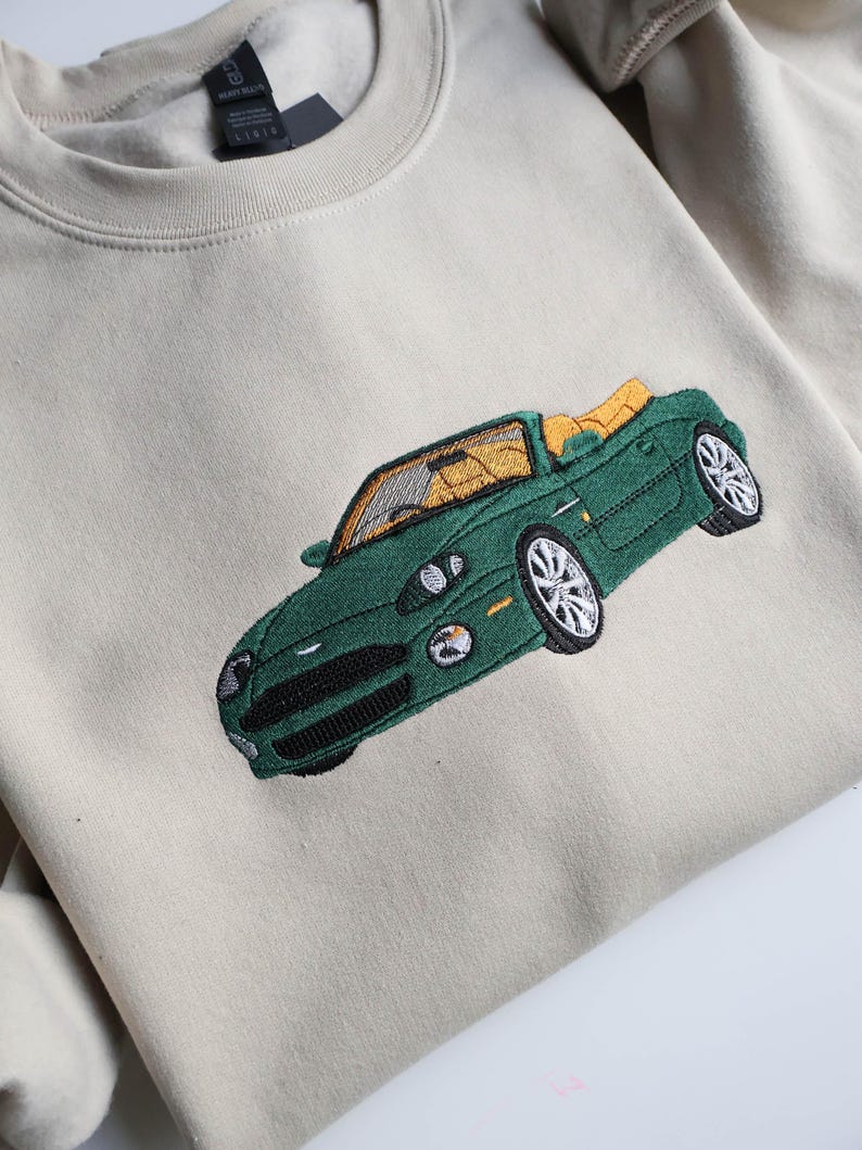May include: A cream-coloured sweatshirt with an embroidered green convertible car design. The car has a yellow interior and detailed stitching. The sweatshirt has a crew neck and a tag on the inside.