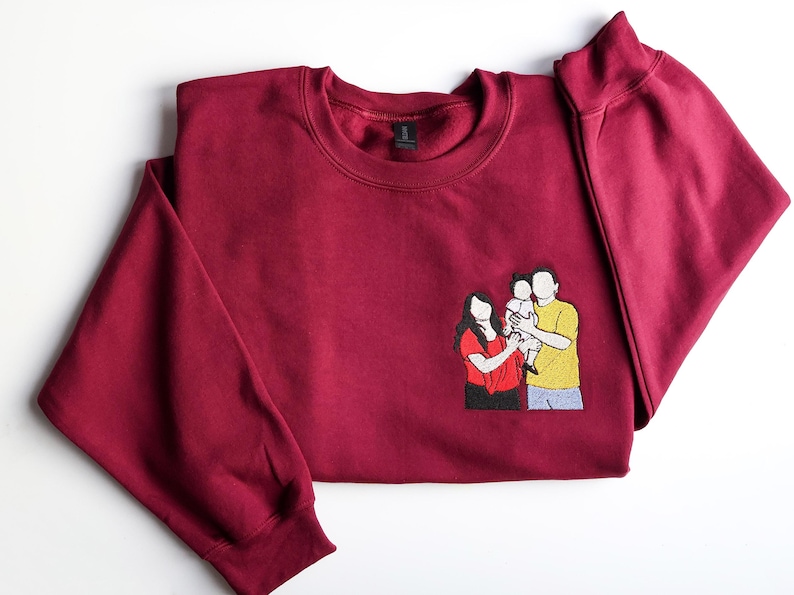 Puede incluir: Una sudadera burdeos con cuello redondo y mangas largas. Un parche bordado en la parte delantera representa a una familia de tres. La familia est&aacute; compuesta por una mujer con camisa roja, un hombre con camisa amarilla y un ni&ntilde;o.